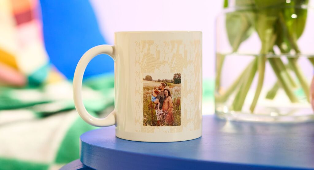 Das Bild zeigt eine weiße Tasse, die auf einem runden blauen Tisch steht. Auf der Tasse ist ein Foto mit einer Familie gedruckt.