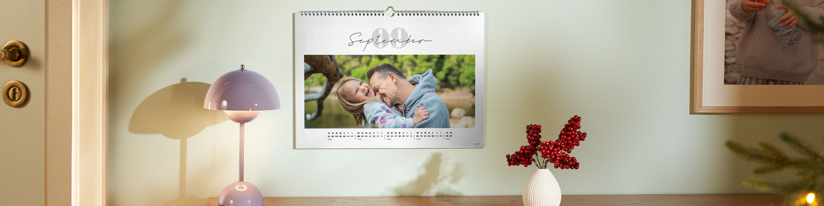 Ein CEWE Fotokalender zeigt ein liebevolles Foto von Vater und Kind in einer warmen Umarmung, über einer Kommode hängend.