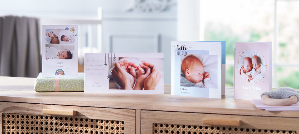 Zwei Hände halten einen Neugeborenen, umgeben von Grußkarten mit einem Baby-Motiv.