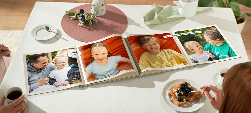 Ein CEWE FOTOBUCH auf einem Tisch mit Essen und einer Pflanze, geöffnet mit Fotos von einer Familie.