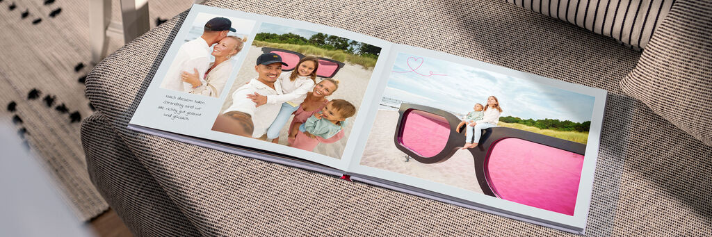 Auf einem Sofa liegt ein Fotobuch. Die Fotos zeigen eine Familie am Strand neben einer überdimensionalen Sonnenbrille mit pinken Gläsern.
