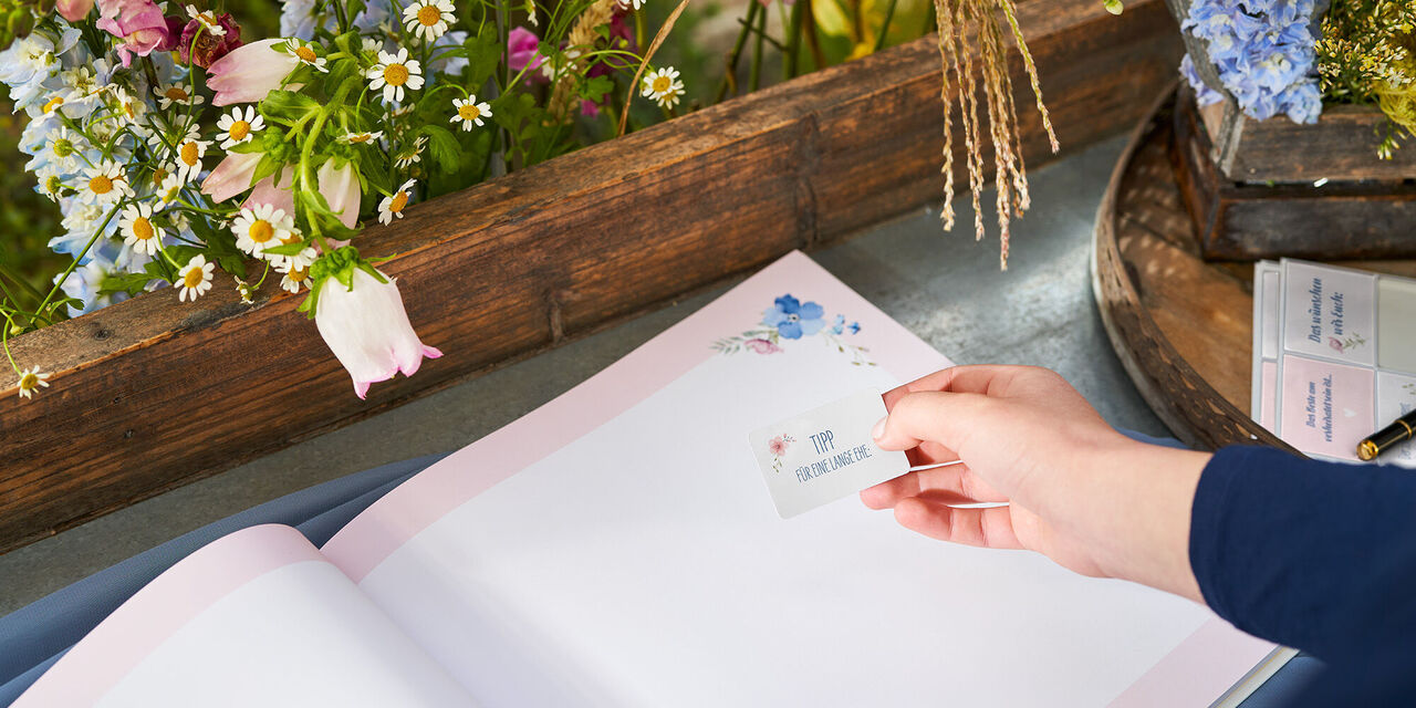 Eine Person hält eine Karte über einen offenen Fotobuch-Hintergrund mit Blumen.