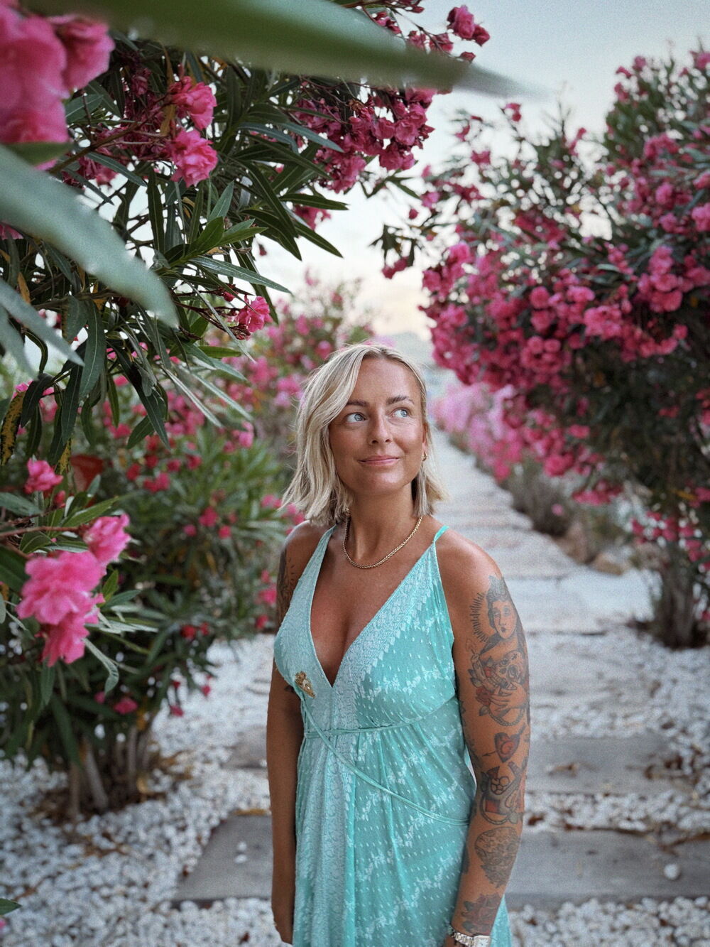 Frau in türkisem Sommerkleid steht zwischen blühenden pinken Oleanderbüschen auf einem steinigen Weg.