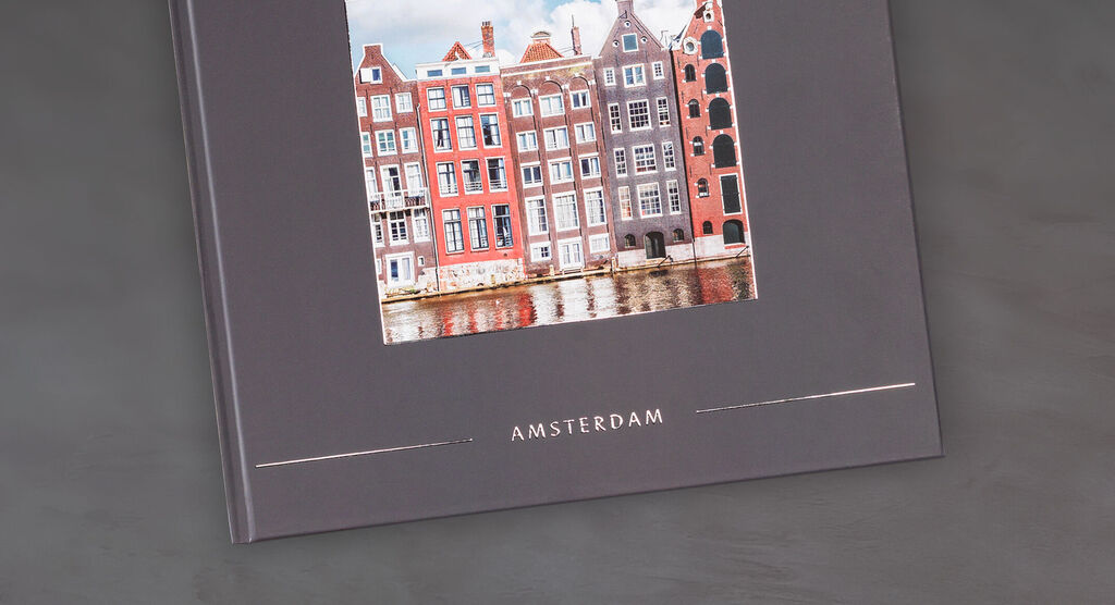 Ein CEWE FOTOBUCH mit einem schönen Foto von bunten Häusern in Amsterdam auf einem eleganten, grauen Hintergrund.