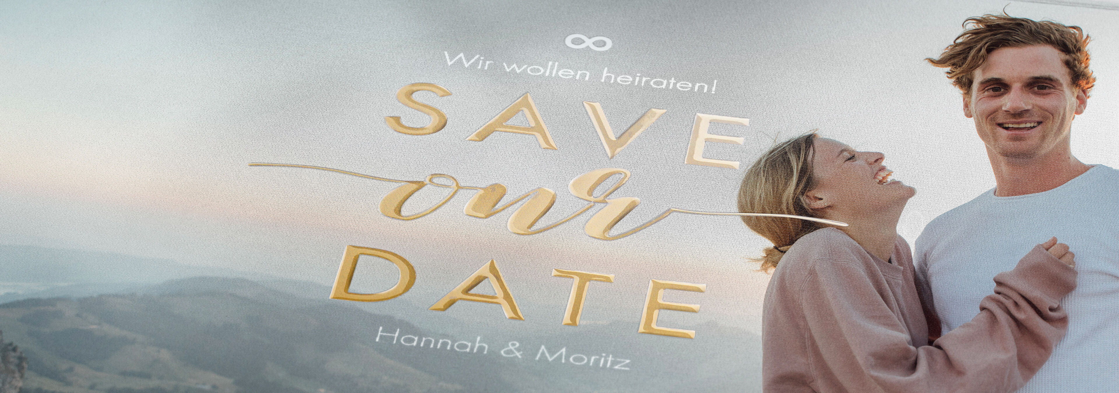 Ein Brautpaar in einer romantischen Umarmung vor einer malerischen Landschaft. Der Text "SAVE our DATE" ist floral gestaltet, darunter stehen die Namen Hannah und Moritz.