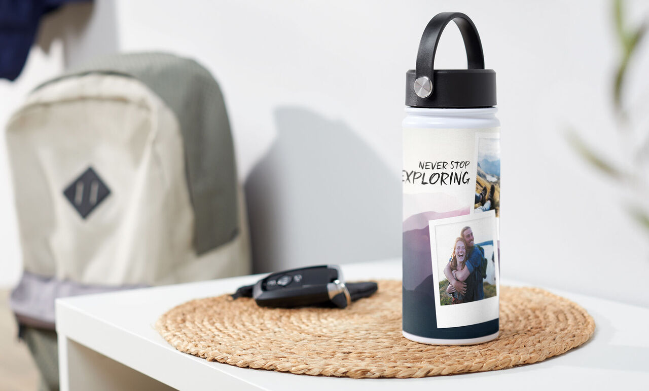 Wasserflasche mit dem Text „Never Stop Exploring“ und Fotos auf einem Tisch. Im Hintergrund ein Rucksack und ein Autoschlüssel.