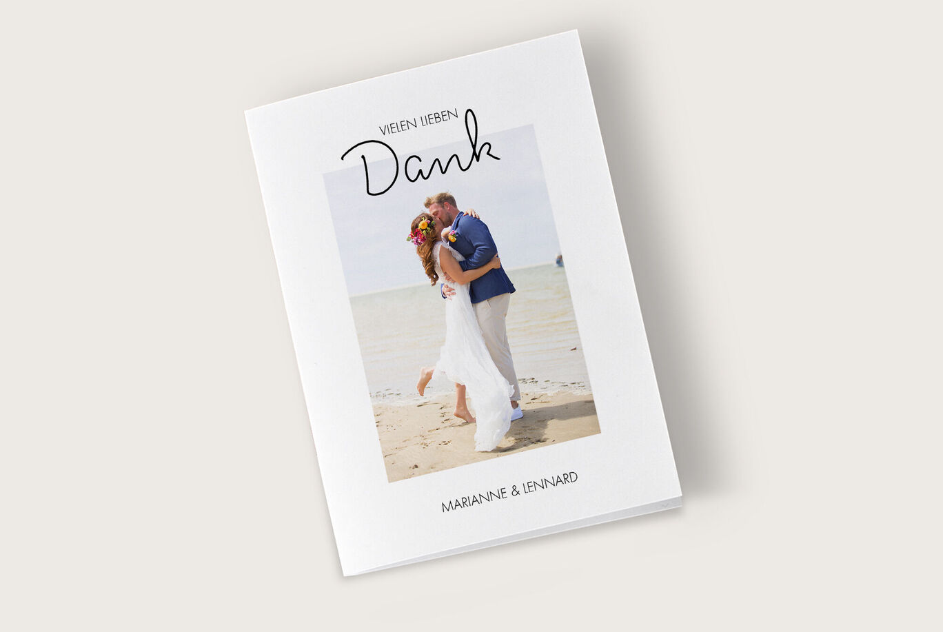 Ein Paar umarmt sich am Strand, während sie Dankbarkeit mit einem Fotobuch ausdrücken. Der Hintergrund zeigt das Meer und den sandigen Strand auf einem hellen Cover.