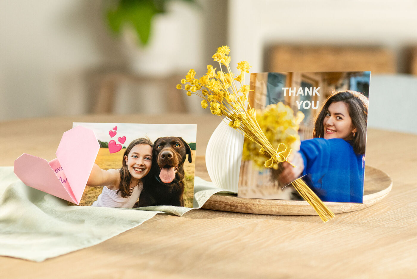 Ein liebevolles Arrangement mit einer CEWE Grußkarte, einem Foto von zwei Freunden, einem Hund, und einer kleinen Vase mit Gelben Blumen auf einem Holztablett.