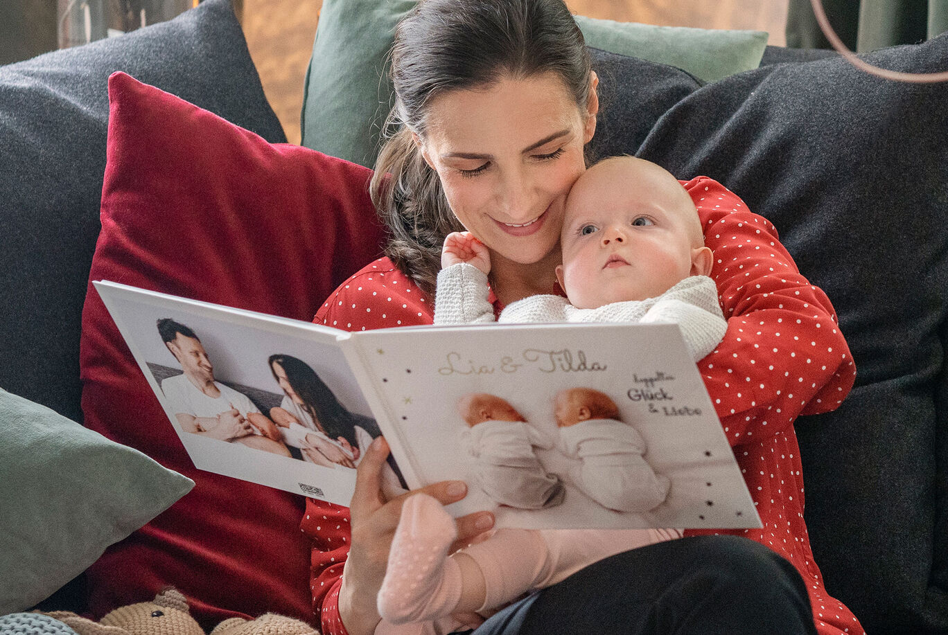 Eine Person liest ein CEWE FOTOBUCH mit einem Baby im Arm, umgeben von gemütlichen Kissen.
