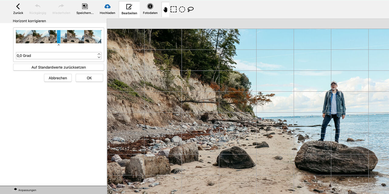 Ein Mann steht auf einem Sandstrand mit großen Felsen. Im Hintergrund sieht man das Meer und eine Freifläche mit Bäumen. Der Himmel ist teilweise bewölkt.
