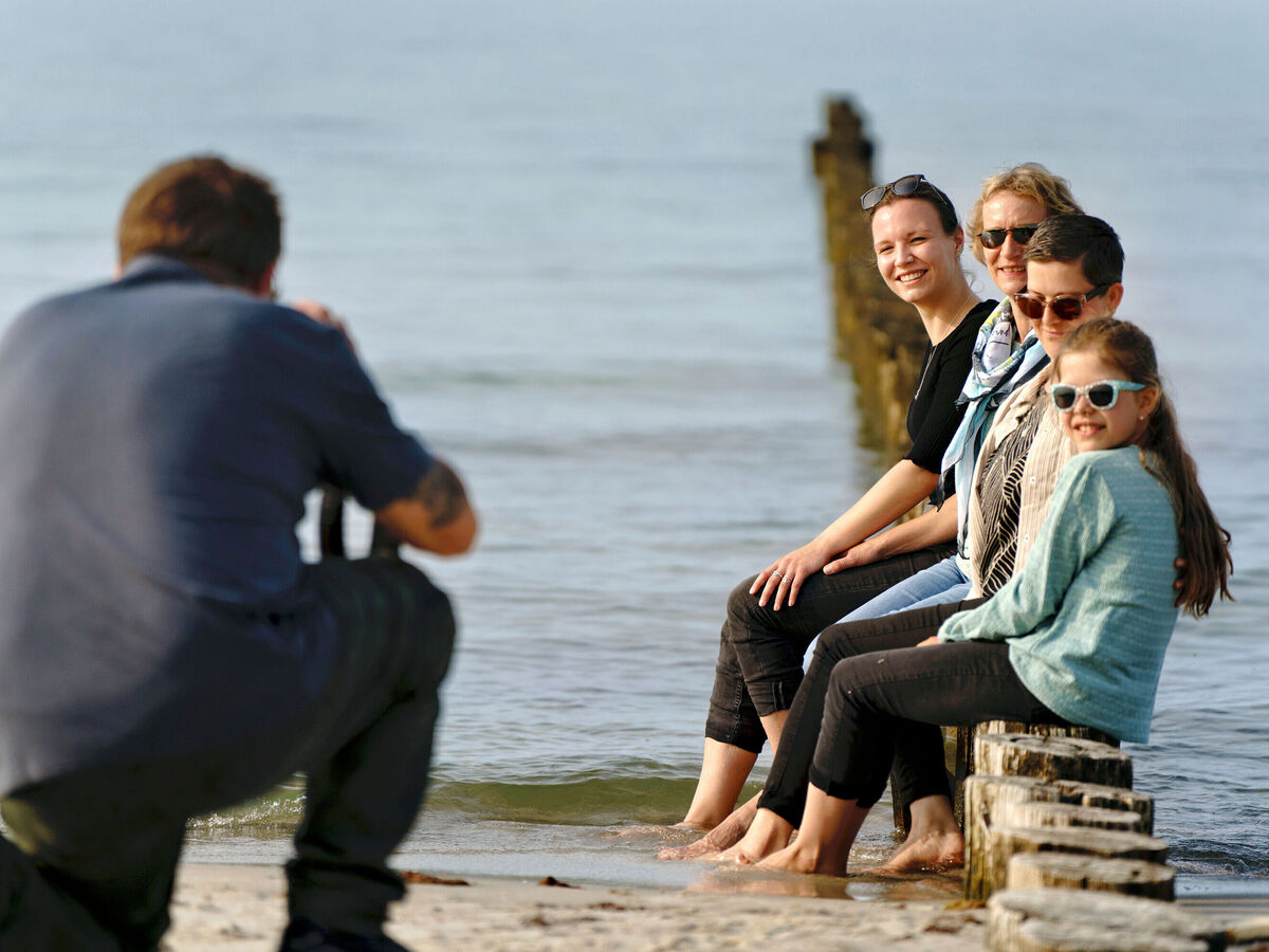 Vier Menschen sitzen lachend auf Holzpfählern an einem Strand und werden von einem Fotografen fotografiert.