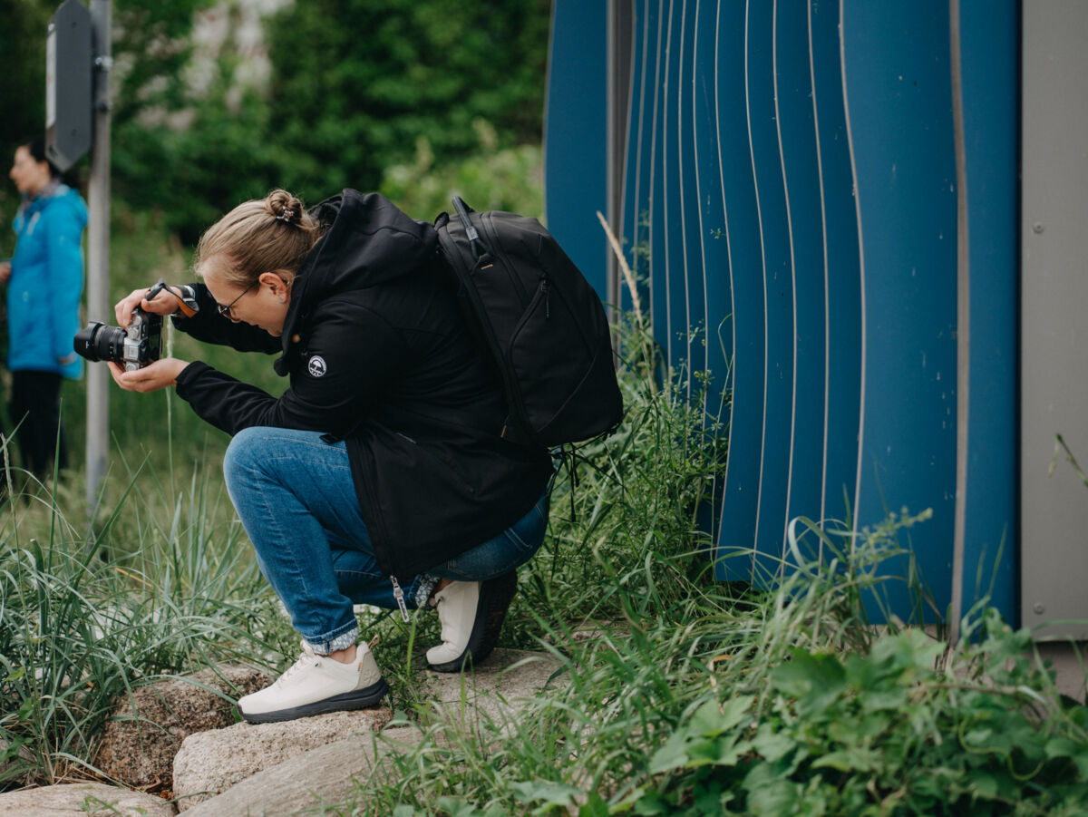 Person hockt im Gras neben einer blauen Wand und fotografiert mit einer Kamera ein Motiv am Boden.