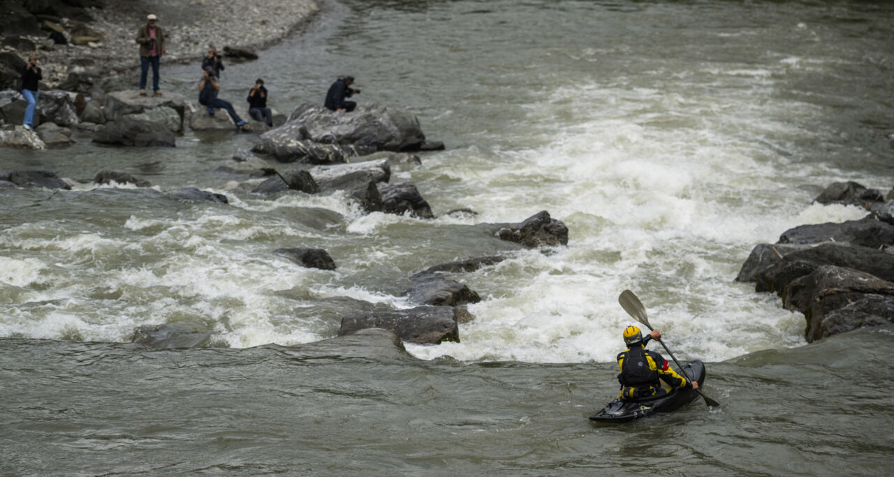Eine Person fährt in einem Kajak auf einem Fluss. Direkt vor ihr liegt eine enge Flussstelle. Im Hintergrund warten Personen mit Kameras.