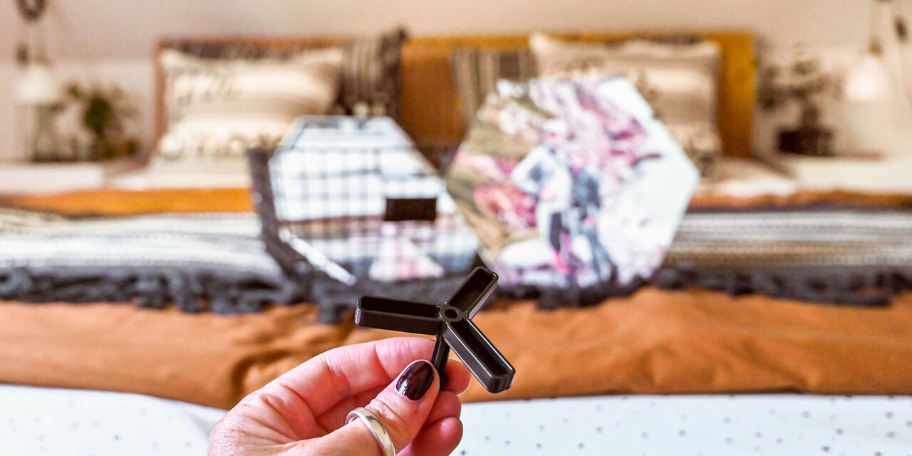 Eine Person hält ein schwarzes Kunststoffteil in der Hand und zeigt darauf. Im Hintergrund sind zwei dekorative Kissen auf einem Bett sichtbar.