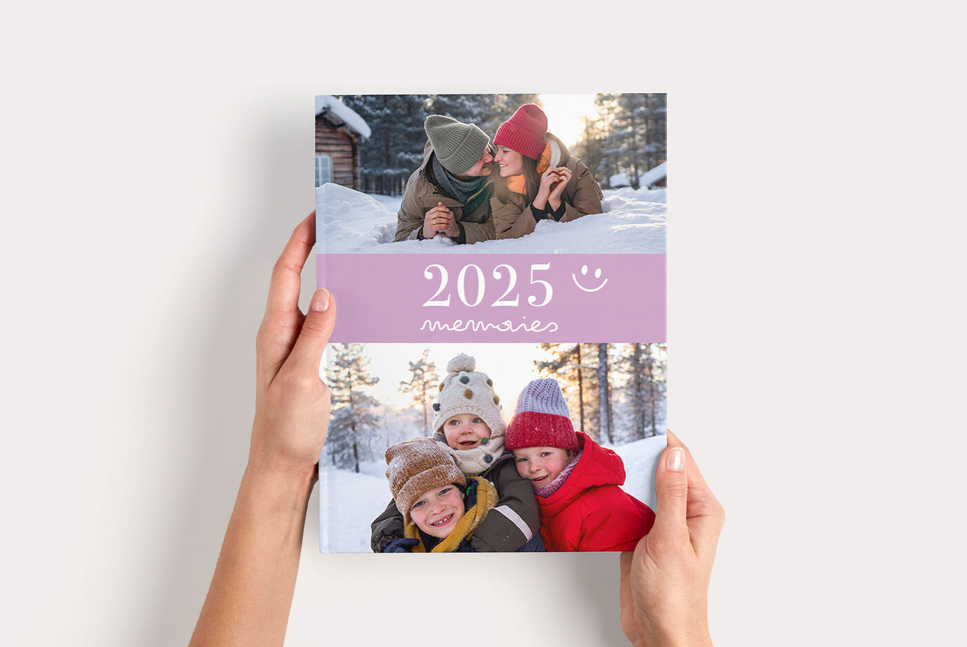 Hände halten ein CEWE FOTOBUCH Groß mit Winterfoto von lachenden Kindern im Schnee und violettem Titelband