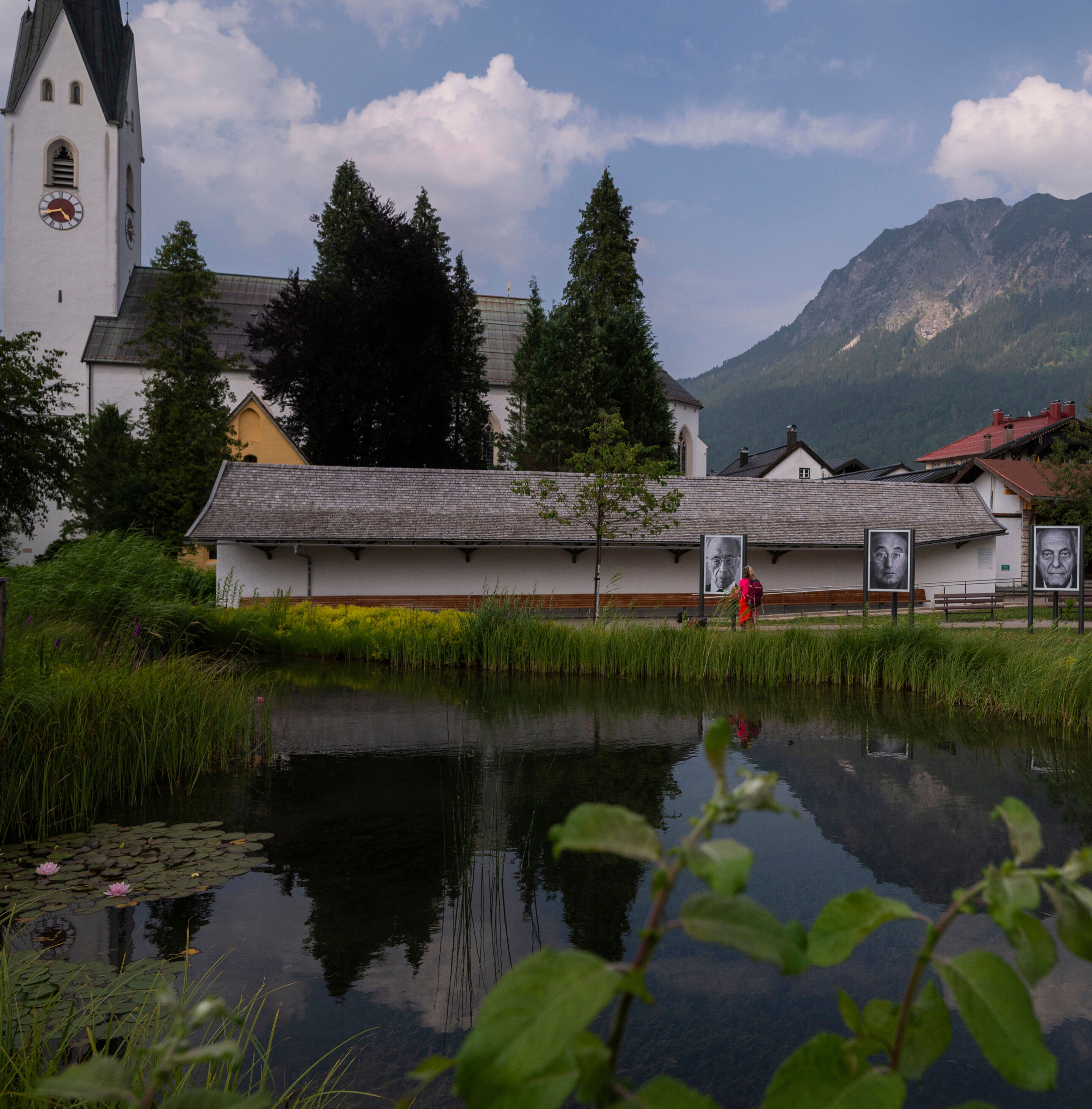 Auf einer grünen Wiese stehen drei große Schwarz-weiß-Fotografien von verschiedenen Menschen. Im Hintergrund sind eine Kirche und ein Berg.