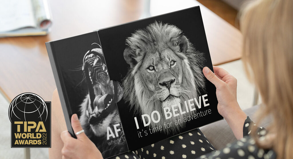 Eine Person hält ein schwarzes Fotobuch mit einem Löwenbild und dem Titel "I DO BELIEVE" in den Händen. Auf dem Tisch liegt ein weiteres Buch.