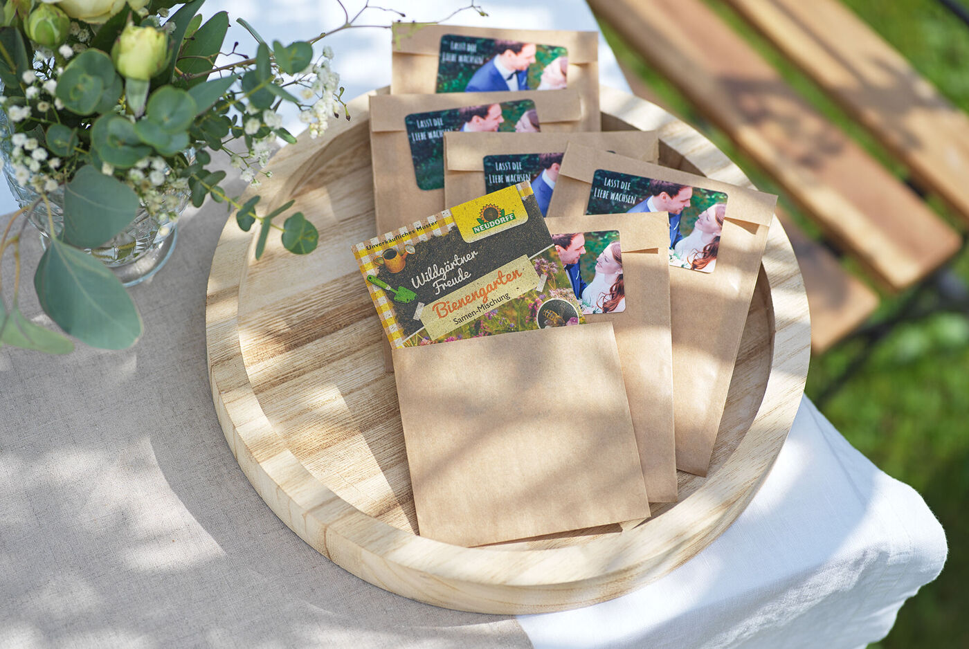 Verschiedene Umschläge mit Samenpäckchen auf einer Holzplatte, umgeben von Pflanzen. Ideal für Gartenliebhaber.