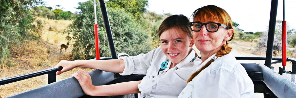 Zwei Personen auf Safari im offenen Fahrzeug mit sanftem Landschaftshintergrund. Ein Tier ist in der Ferne zu sehen.