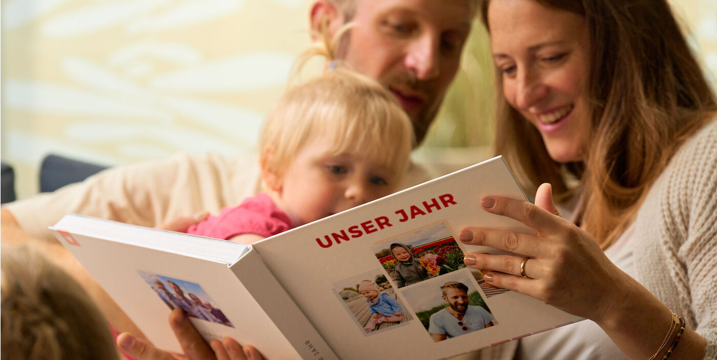 Eine Familie sitzt beisammen und betrachtet ein geöffnetes CEWE FOTOBUCH mit mehreren Familienbildern auf dem Einband.