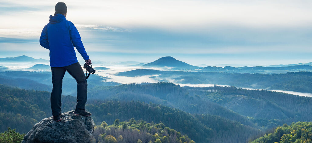 Mann in blauer Jacke steht auf einem Felsen und schaut auf eine bewaldete Berglandschaft.