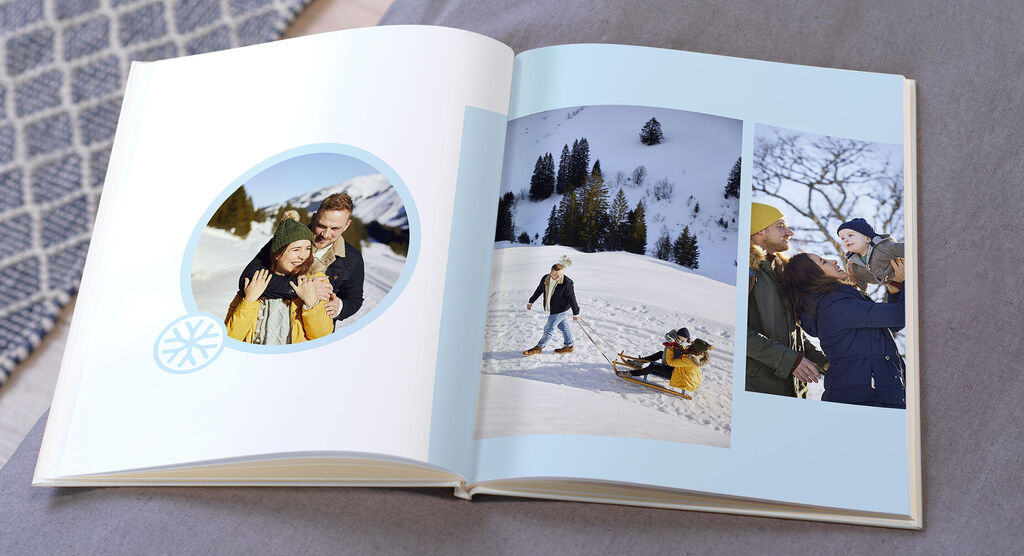 Zwei Seiten eines offenen Fotobuchs mit Urlaubserinnerungen in den Bergen. Eine Seite zeigt ein großes Foto von einer Umarmung im Schnee, die andere zeigt drei kleinere Fotos von Freunden, die im Schnee Spaß haben, und einer Person, die lächelnd posier