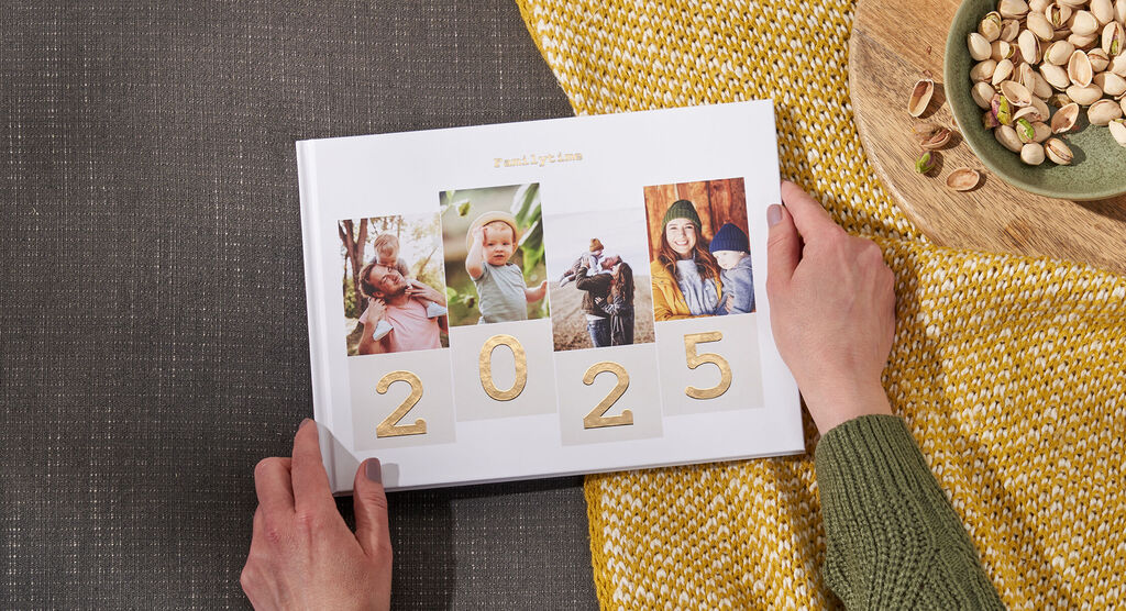 Ein offenes Fotobuch mit Bildern aus dem Jahr 2024 auf einem Tisch. Die Hände einer Person halten das Buch, daneben liegen Pistazien auf einem Teller.