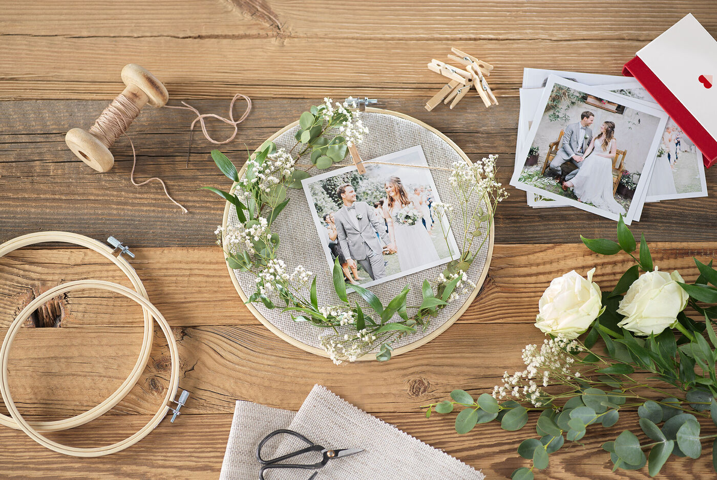 Ein liebevoll gestalteter Tisch mit Hochzeitsfotos, einer Blütenkranz-Dekoration, Scheren und Stoff, ideal für kreative Erinnerungen.