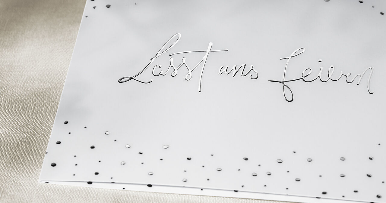 Elegante Grußkarte mit der Aufschrift 'Lasst uns feiern' in geschwungener Schrift, verziert mit kleinen Punkten.