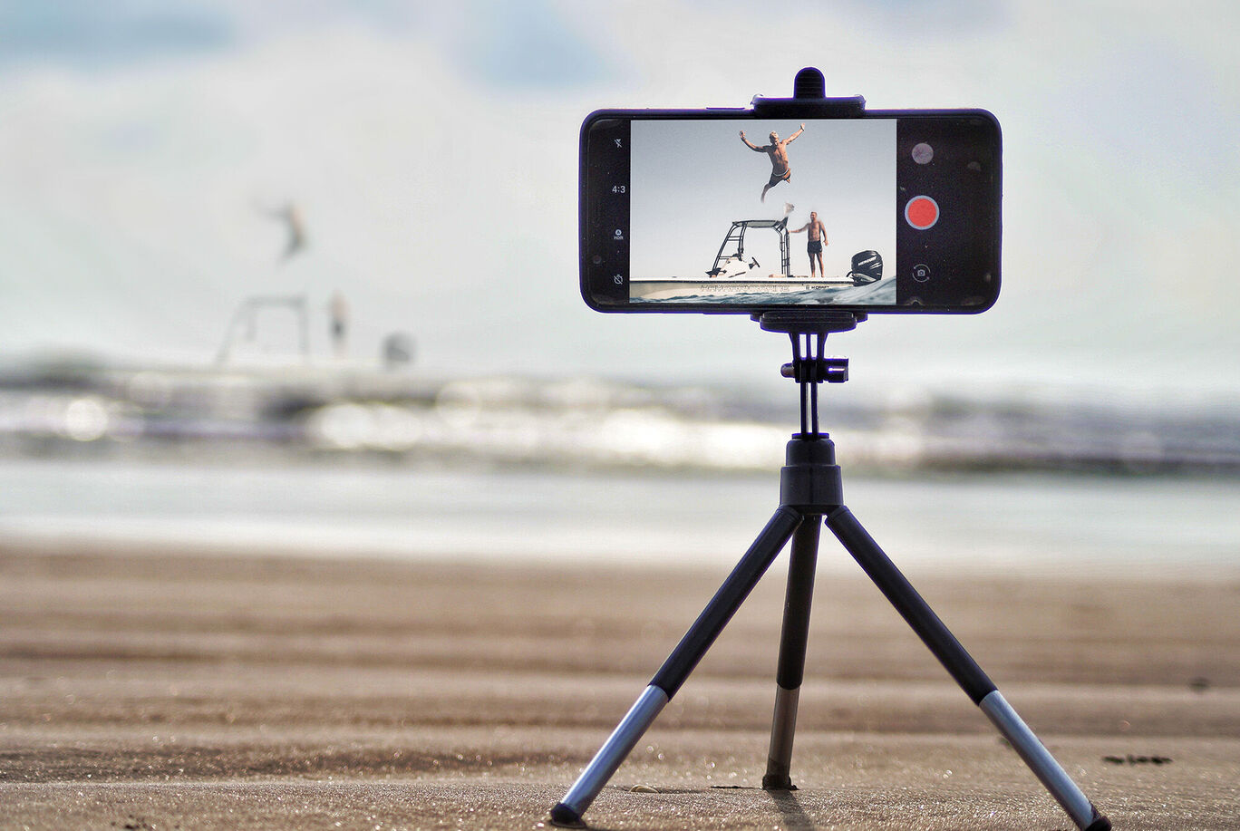 Ein Smartphone auf einem Stativ filmt einen Menschen, der am Strand springt und lächelt