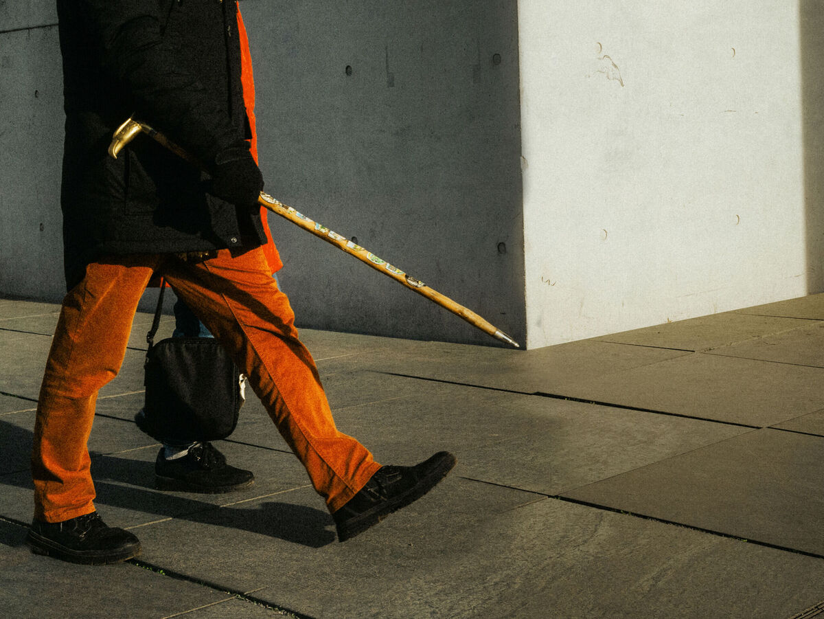 Person mit orangefarbener Hose geht über einen hellen Gehweg und hält einen langen Stock in der Hand