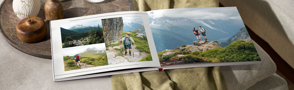Ein CEWE FOTOBUCH zeigt Urlaubsfotos von zwei Wanderern auf einem Felsen in den Bergen. Sie lachen und genießen die Aussicht auf die schöne Landschaft.