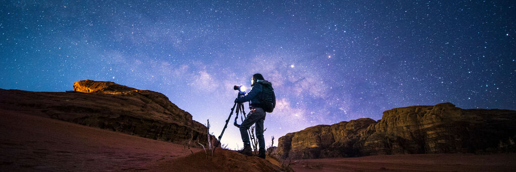 Mann steht auf einer sandigen Fläche und fotografiert den Nachtsternenhimmel.