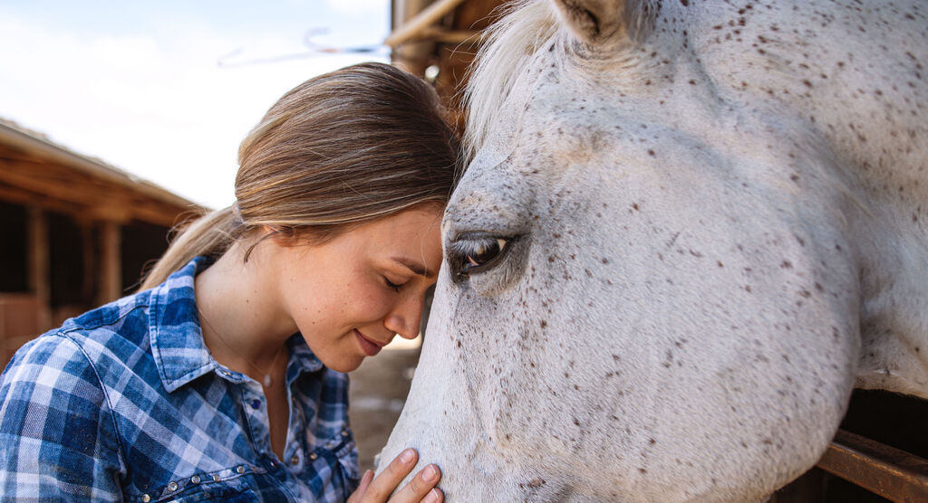 Eine Frau mit blonden Haaren lehnt sich sanft an ein weißes Pferd, umgeben von einem landwirtschaftlichen Hintergrund