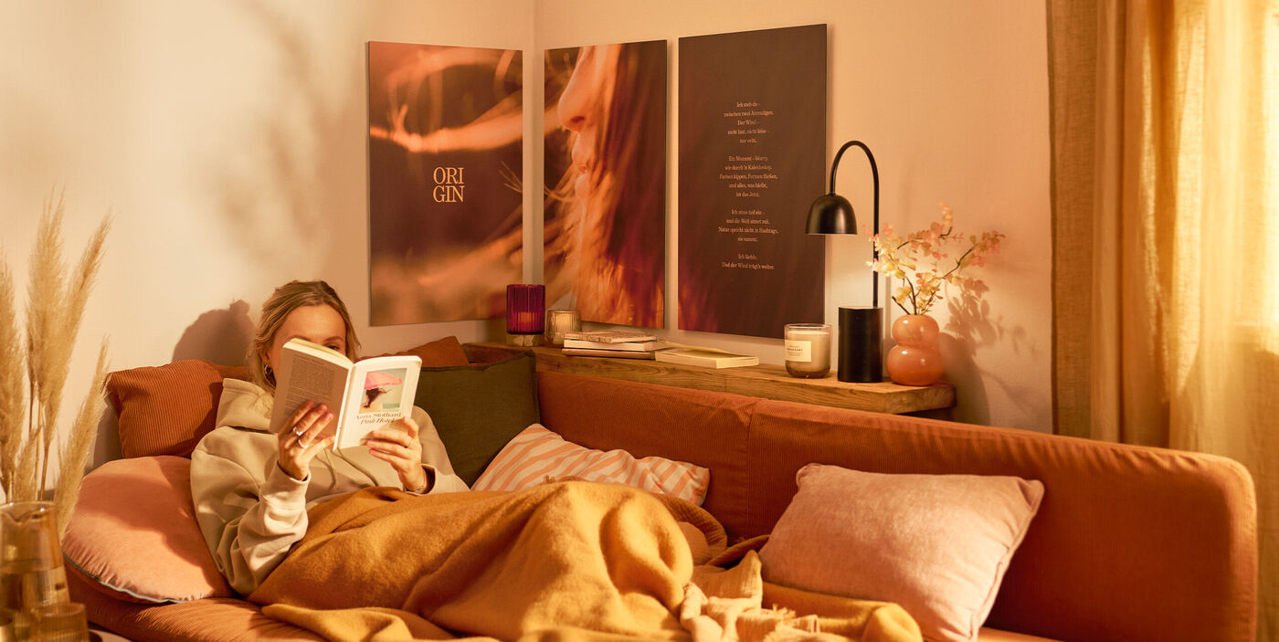Eine Frau sitzt auf einem Sofa und liest ein Buch. Über ihr hängt ein mehrteiliges Wandbild.