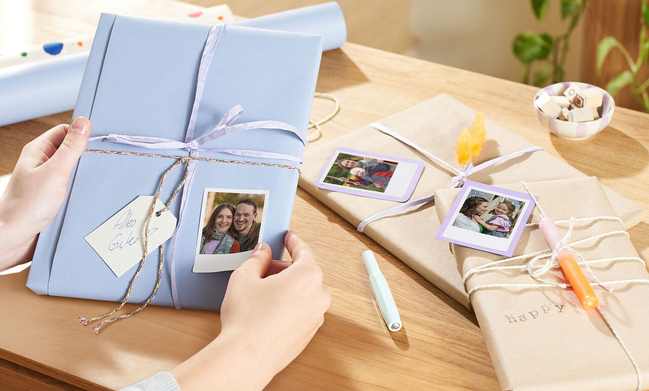 Eine Person hält ein kreativ verpacktes Geschenk mit Polaroid-Fotos darauf. Auf dem Tisch liegen weitere liebevoll verpackte Geschenke.