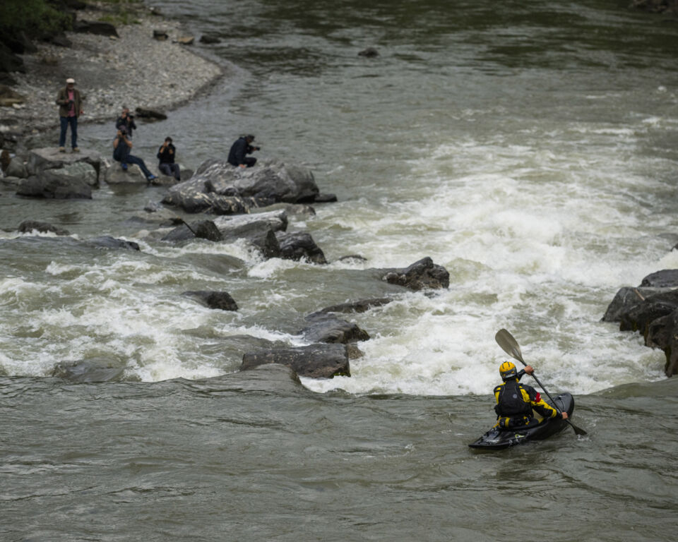 Eine Person fährt in einem Kajak auf einem Fluss. Direkt vor ihr liegt eine enge Flussstelle. Im Hintergrund warten Personen mit Kameras.