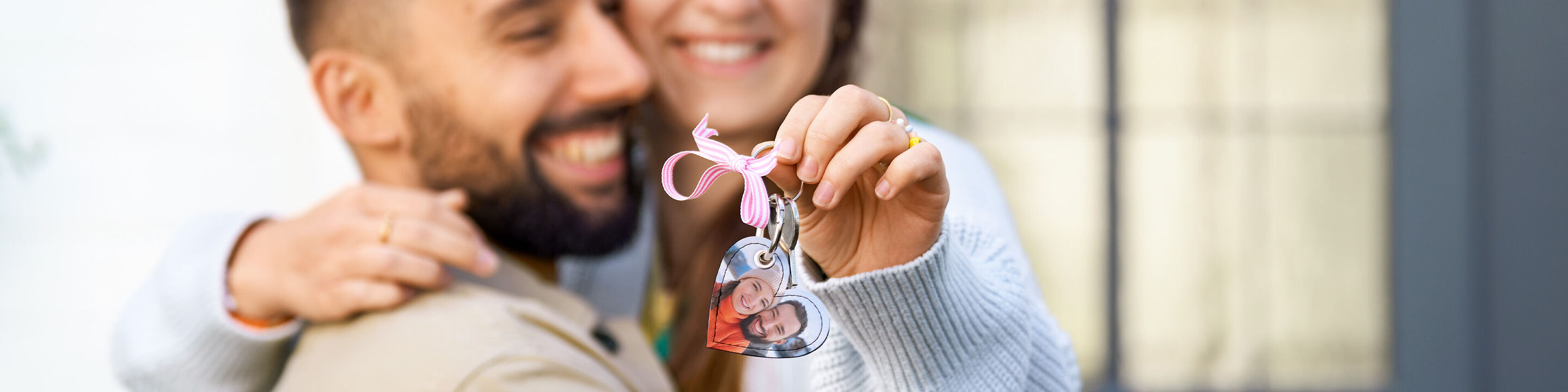 Lächelndes Paar, Person hält einen herzförmigen Schlüsselanhänger mit Foto und rosa Schleife