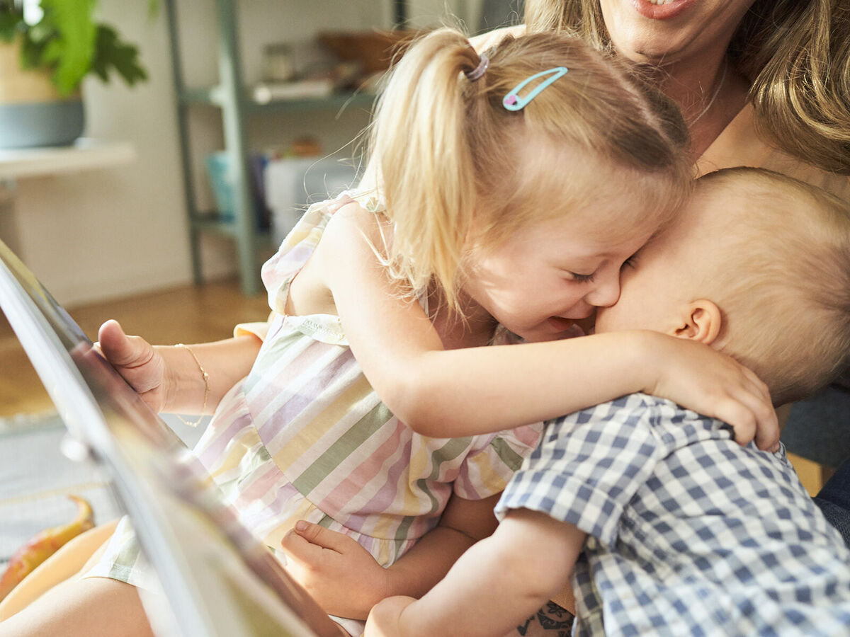 Ein fröhliches Kind umarmt ein Baby bei einer gemütlichen Lesezeit mit ihrer liebevollen Mutter im Hintergrund