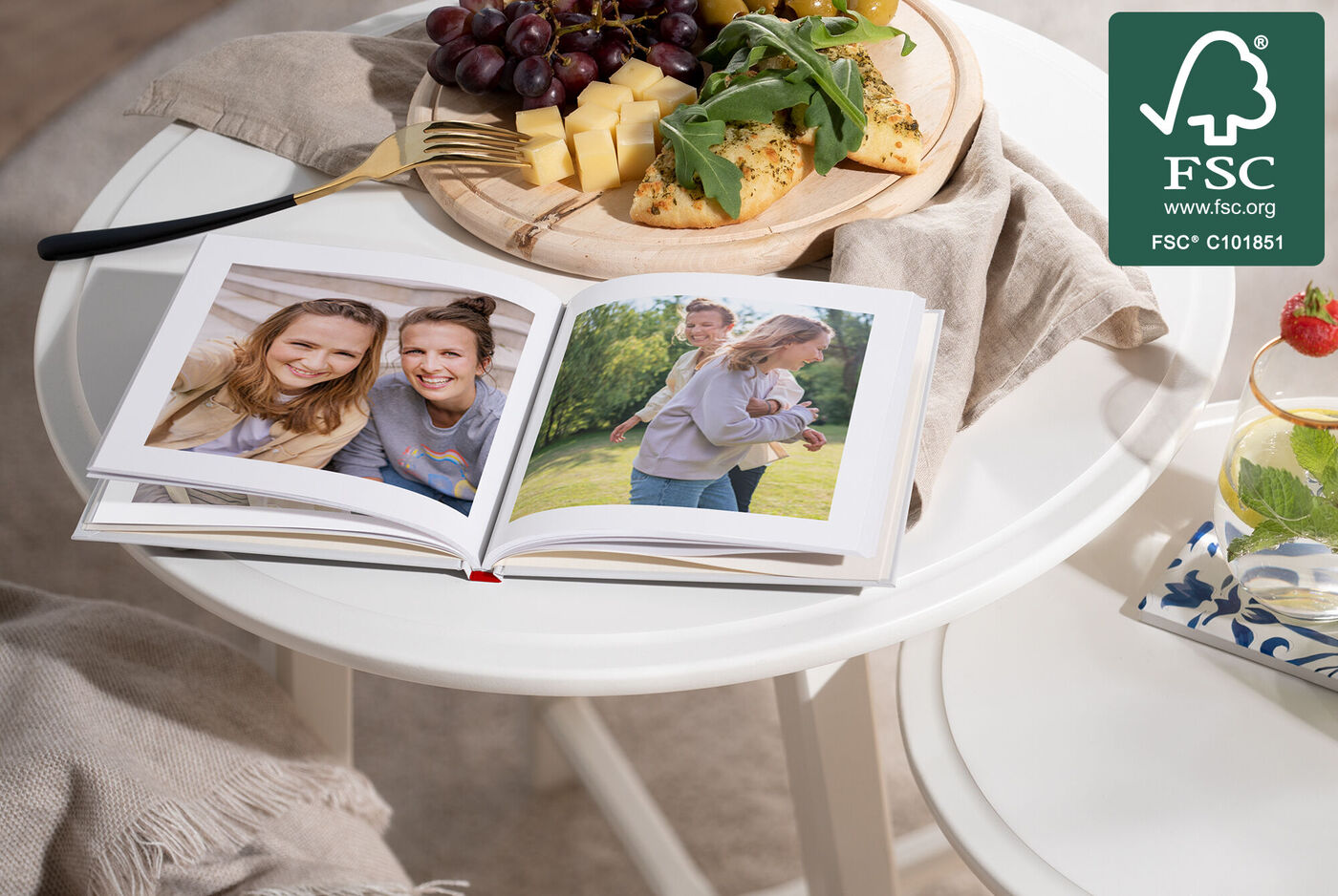 Ein offenes Fotobuch mit zwei Bildern auf einem Tisch, umgeben von Früchten und Käse.