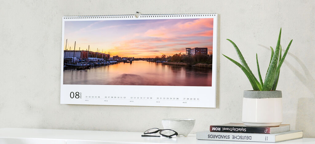 Ein wunderschöner Kalender hängt an der Wand und zeigt einen lebhaften Sonnenuntergang über einem ruhigen Hafen. Der Monat zeigt August mit klaren Zahlen.