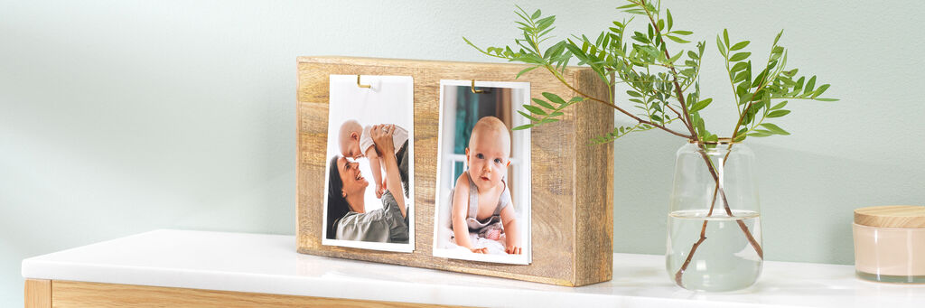 Zwei Fotos von Kindern in einem Bilderrahmen auf einer schlichten Holzablage neben einer Pflanze in einem Glas.