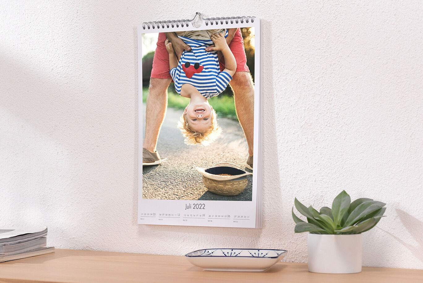 Ein kreativer Fotokalender für Juli 2022, zeigt einen Jungen, der kopfüber hängt, während seine Eltern nebeneinander stehen. Der Junge lacht voller Freude.