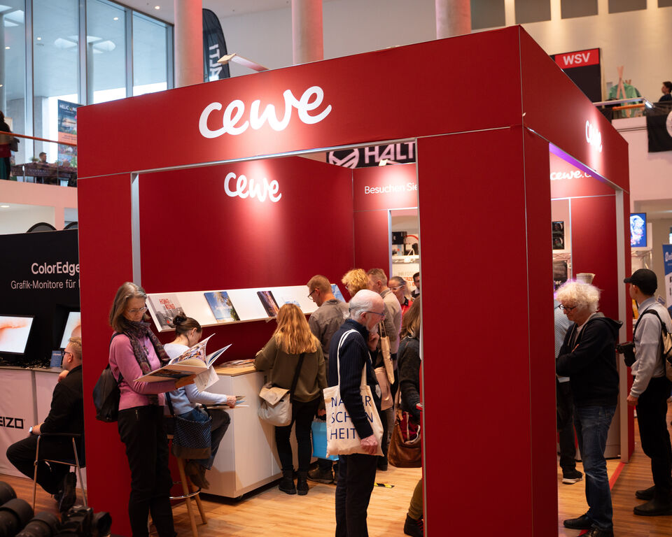 In einer Halle ist ein Messestand von CEWE aufgebaut. Viele Leute betrachten den Stand und die ausgelegten Fotobücher.