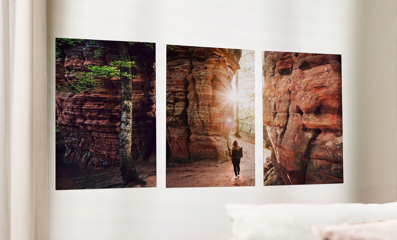 Drei beeindruckende Fotodrucke in warmen Erdtönen an einer Wand, mit Sonnenstrahlen, die in die Szene scheinen. Der mittlere Druck zeigt eine Person, die einen Pfad entlang geht.