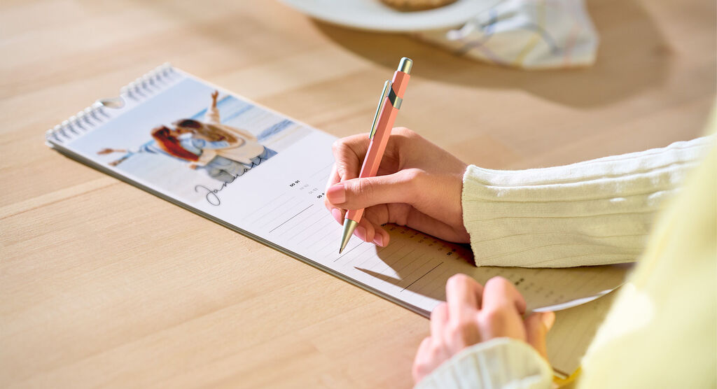 Person schreibt mit rosa Stift in CEWE Fotokalender mit Strandmotiv