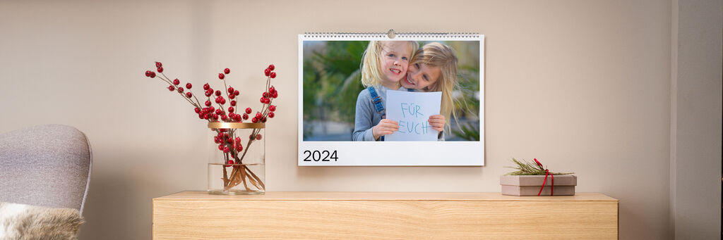 Ein Kalender 2024 steht auf einem Tisch. Eine Person hält ein Blatt Papier mit einer Zeichnung. Im Hintergrund sieht man eine Pflanze.