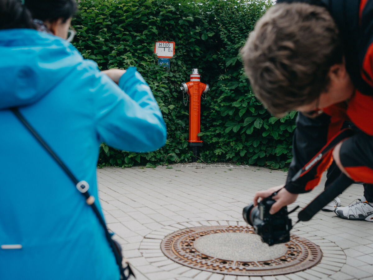 Zwei Personen fotografieren einen Kanaldeckel auf einem Gehweg, im Hintergrund steht ein roter Hydrant vor einer grünen Hecke.