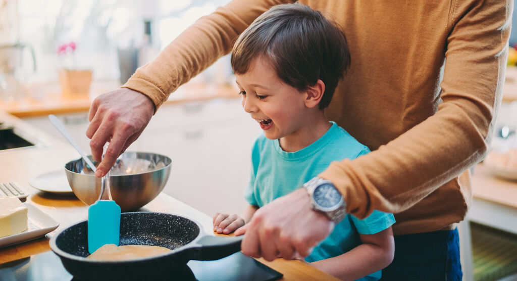 Ein Erwachsener und ein Kind backen zusammen in der Küche. Der Erwachsene hält einen Spatel und wendet einen Pfannkuchen.