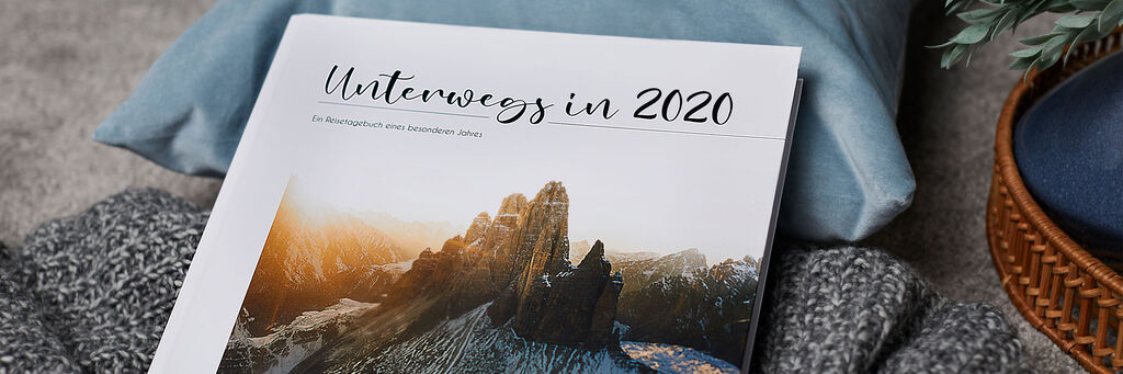 Ein offenes CEWE Fotobuch mit dem Titel „Unterwegs in 2020“. Es liegt auf einem grauen, gestrickten Pullover und zeigt eine atemberaubende Berglandschaft auf der ersten Seite. Die Farben sind warm und einladend.